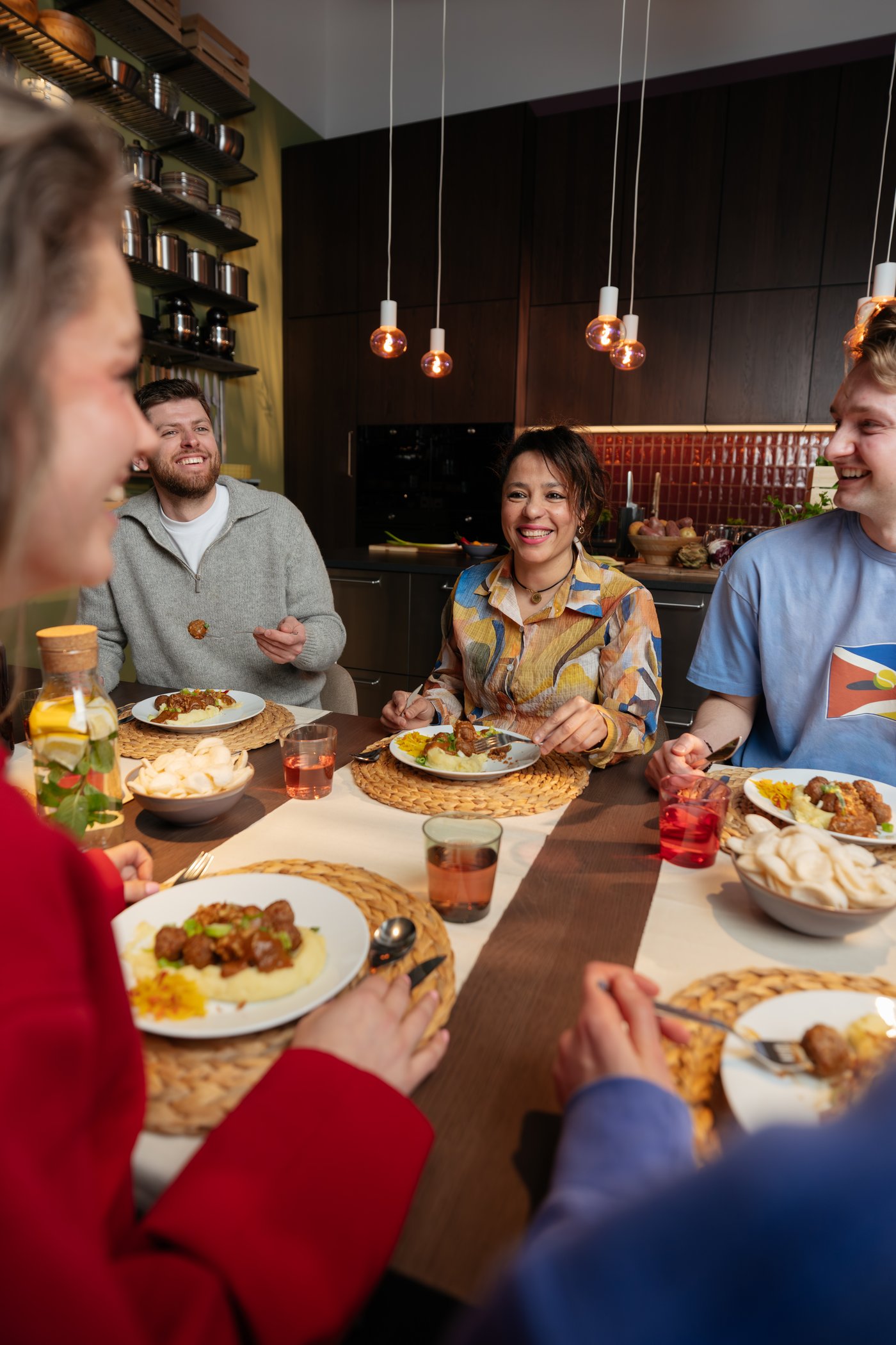 A number of people are sitting at a table. They are smiling at each other. In front of them they have a plate with meatballs. On the table there are bowls of prawn crackers, a bottle of water and glasses filled with lemonade.