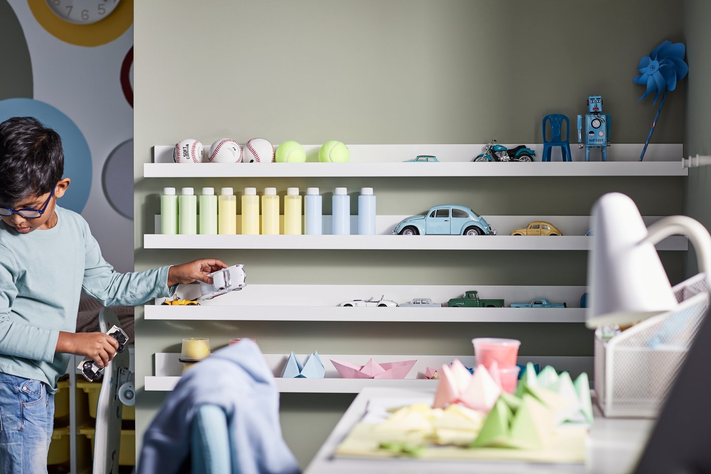 Ett barn leker med leksaksbilar i ett sovrum med vita MOSSLANDA tavelhyllor på väggen med pappersmodeller och flaskor.