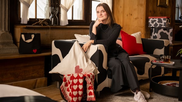 Designerin Lucie De Martin sitzt auf einem KLIPPAN Sofa mit TOSSDAN Bezug in schwarz-weiss. Eine Stofftasche, ein Kissen und Decke mit Herzen und andere Produkte aus der TOSSDAN Kollektion sind zu sehen.