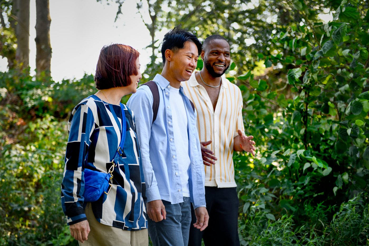 Tre personer går tillsammans i en utomhusmiljö med träd och växter i bakgrunden.
