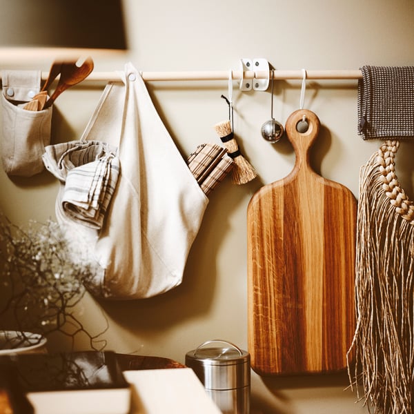 An oak chopping board, cooking utensils, kitchenware and a tea infuser hanging from NEREBY rails on a kitchen wall.