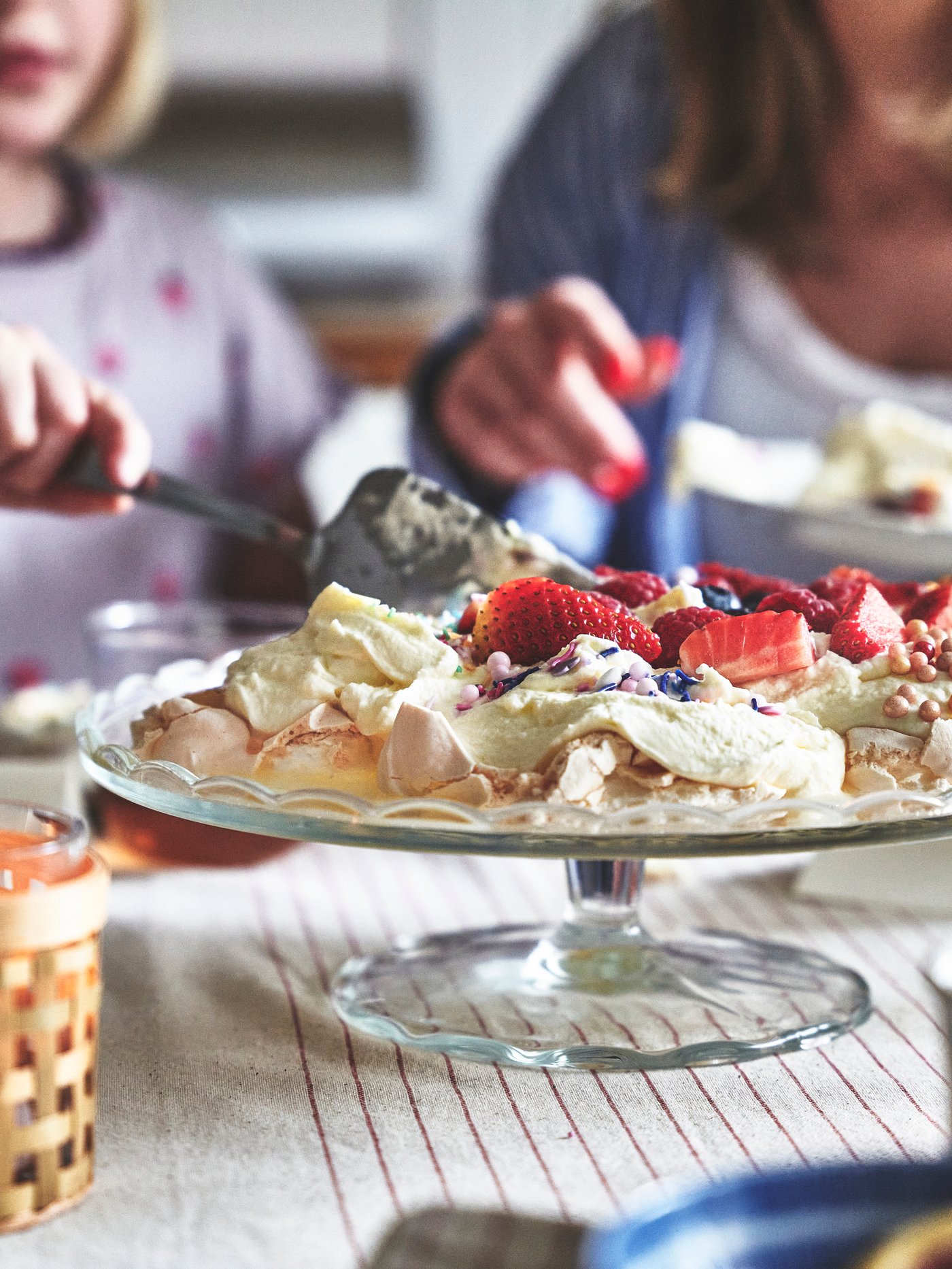 Närbild av ett dukat bord med desserttillbehör, där ett barn häller något i en blåvit skål. På bordet finns glas med dryck, kakor, jordgubbar, vispad grädde och ett litet glas med en blomma.