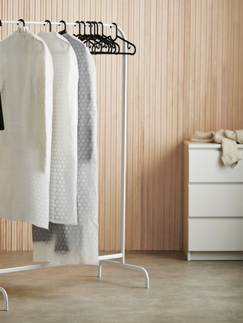 Bedroom with white clothes rack with metal frame, clothes on hangers, black hangers, white chest with three drawers.