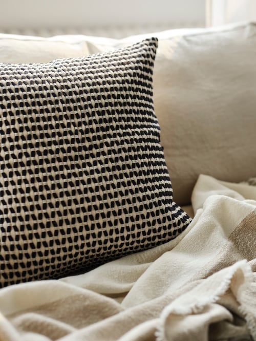 Housse de coussin KUSTFLY à motifs en beige et noir et jeté léger sur un fauteuil BACKSÄLEN.