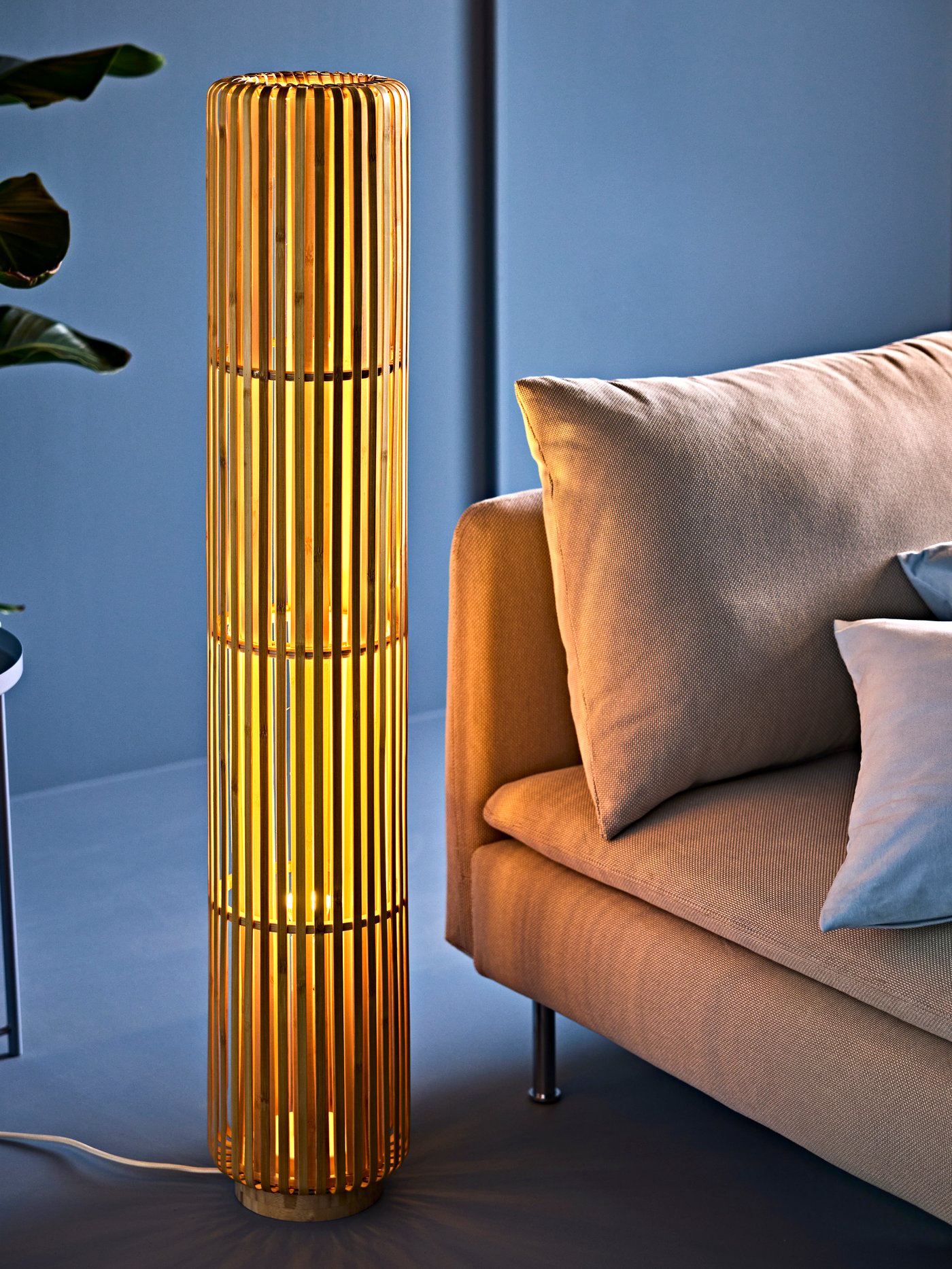 A living room with a VARPTROSS bamboo floor lamp casting a warm glow, beside a beige sofa with cushion and pillows.