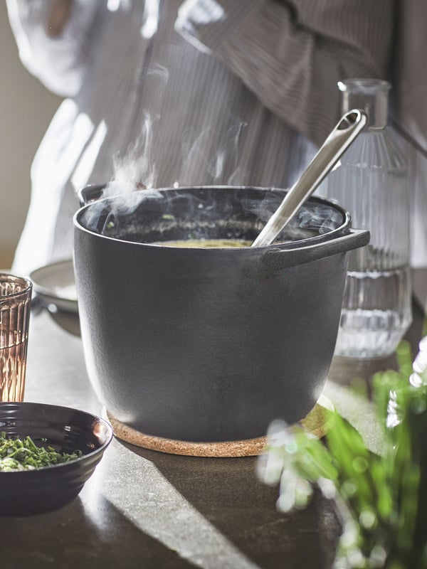A VARDAGEN pot made of matt black enameled cast iron is standing on a pot stand. It has food inside and a ladle sticking out.