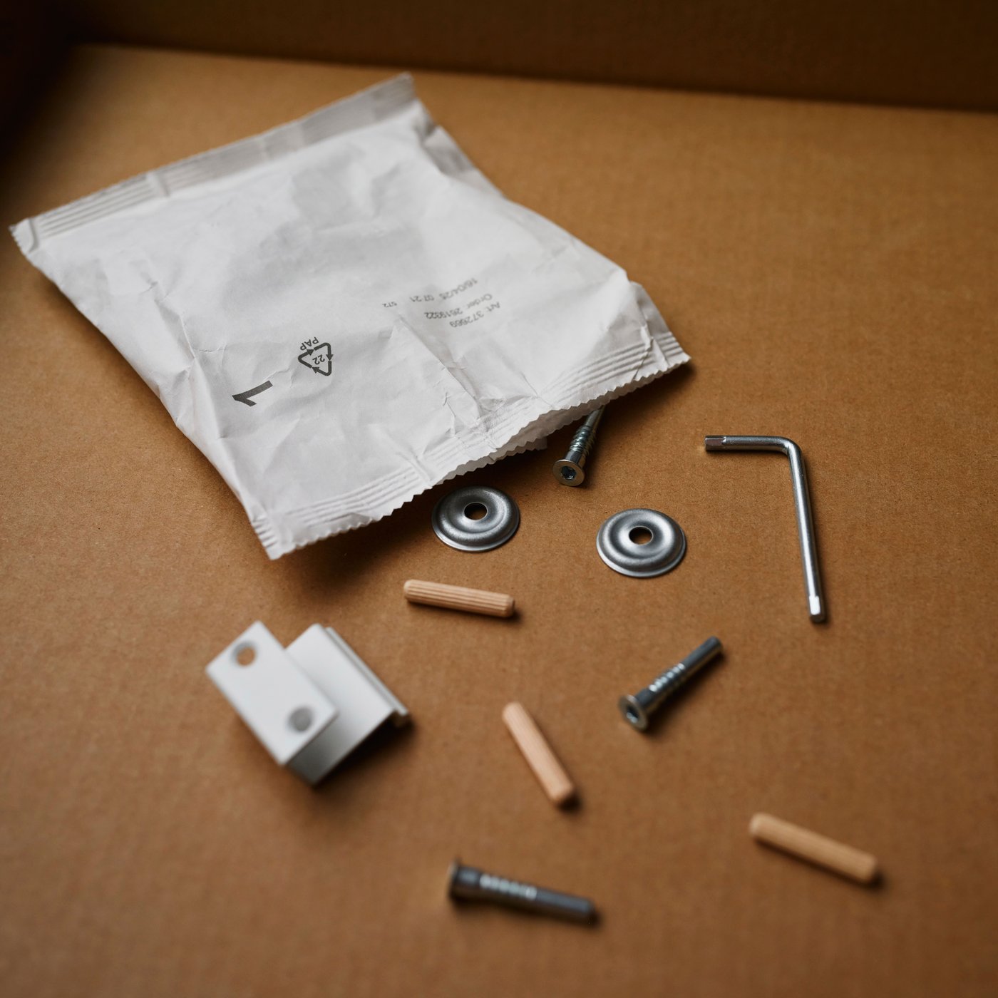 A paper-based fitting bag is on a cardboard surface with dowels, screws, allen keys and other assembly parts nearby.