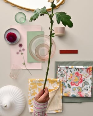 한 여성의 손이 평평한 표면에 놓인 핑크색 종이 한 장 위에 보라색 압화를 놓고 있습니다. 라이트그린 패브릭 한 장과 컬러풀한 플로럴 패턴의 이불커버가 추가되고, 브라운 유리 접시와 다른 액세서리가 더해져 보기 좋은 콜라주가 만들어집니다.