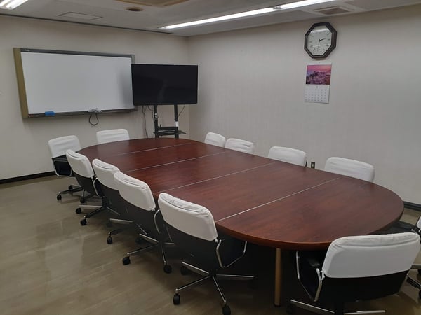 長円形の木製テーブルを囲んで白いオフィスチェアを配置した会議室。壁にホワイトボードと時計が掛かっている。