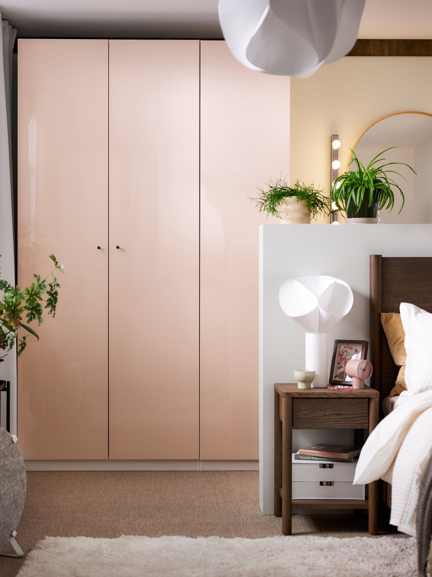 Modernt sovrum med ljusrosa garderob, vit taklampa, vägg med spegel och växter, samt säng med vita sängkläder och ett nattduksbord med lampa och böcker.