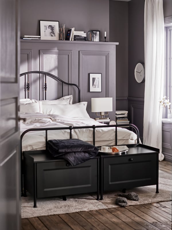 Two black KORNSJÖ storage benches and a bed with a black bed frame made with white/dark gray bed linen on a light gray rug.