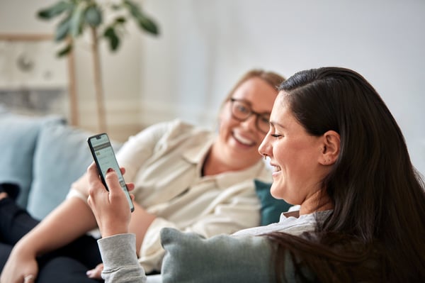 Two women sitting on a sofa smiling, one of them is holding a phone.