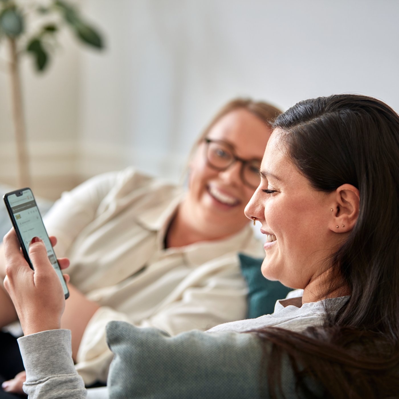 Two women shopping online with mobile phone while sitting on a blue couch.