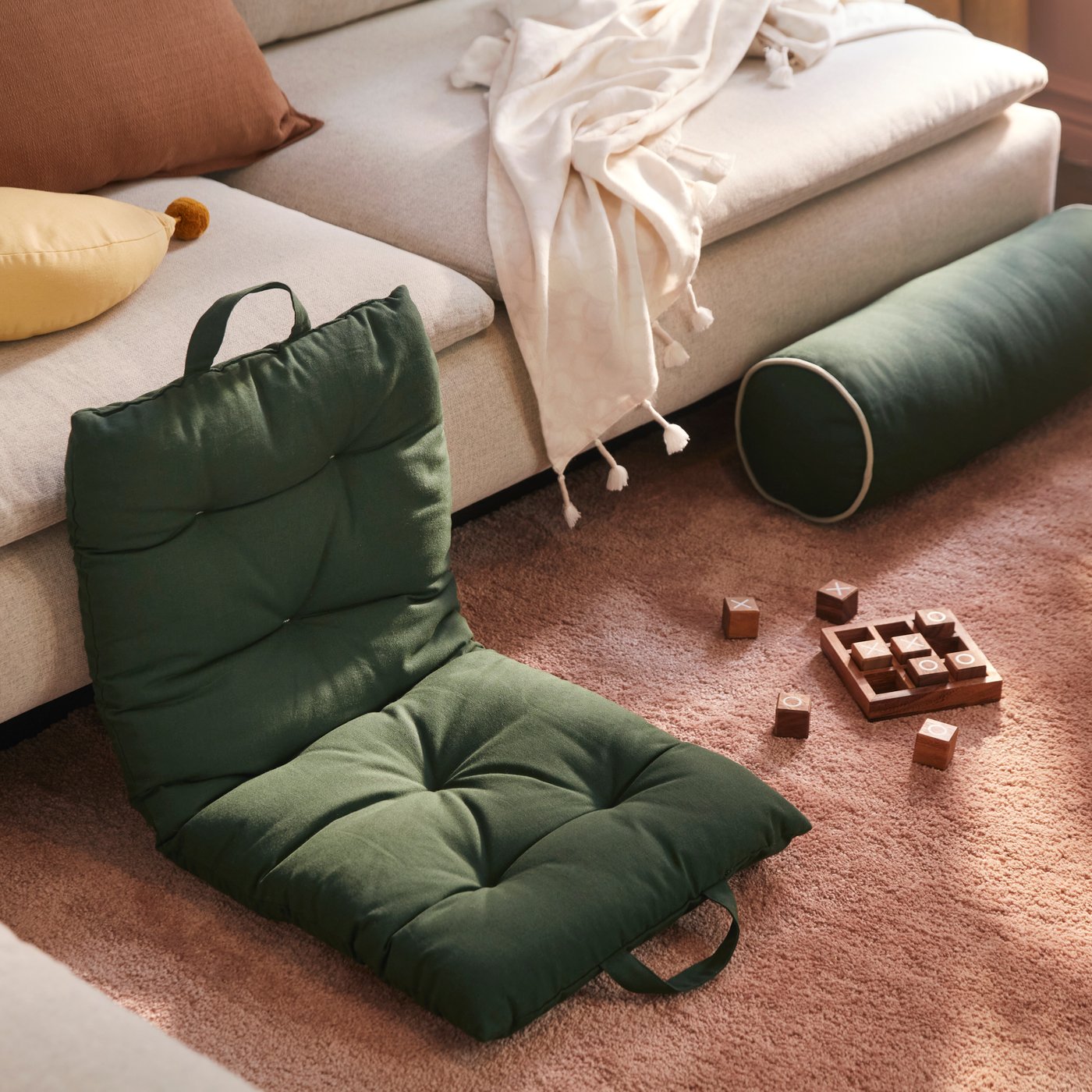 A dark green GULDBRAXEN floor cushion placed on a rug beside a sofa with cushions and blanket.