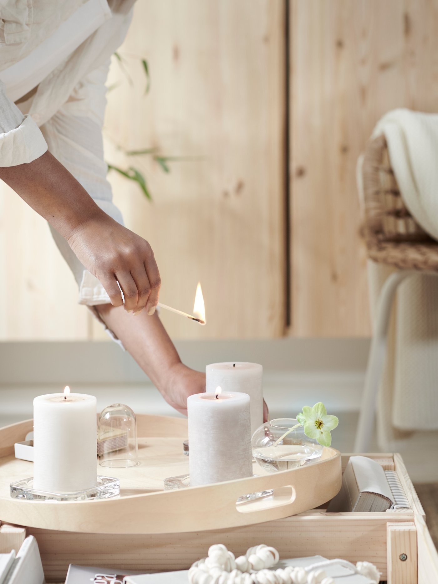 A woman is lighting three LUGGA scented block candles placed on a wooden tray in front of a solid pine IVAR cabinet.