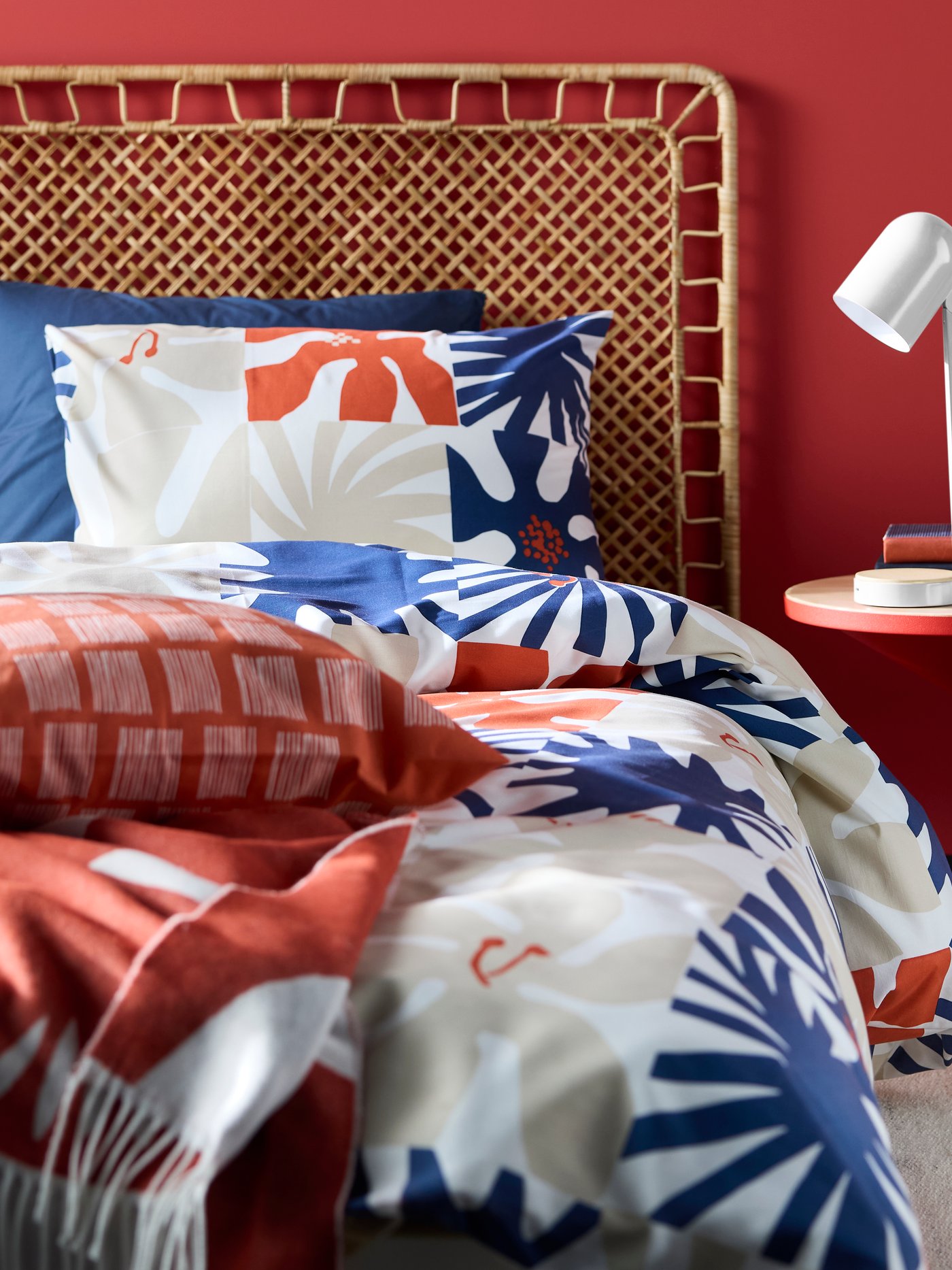 A white desk lamp sitting on a red side table placed beside a bed with a rattan headboard, made with HYLSNEJLIKA bed linen.