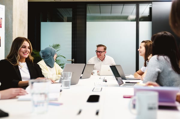 In einem Meetingraum sitzen verschiedenen Personen mit Laptop, Notizblöcken und Wassergläsern an einem Konferenztisch.