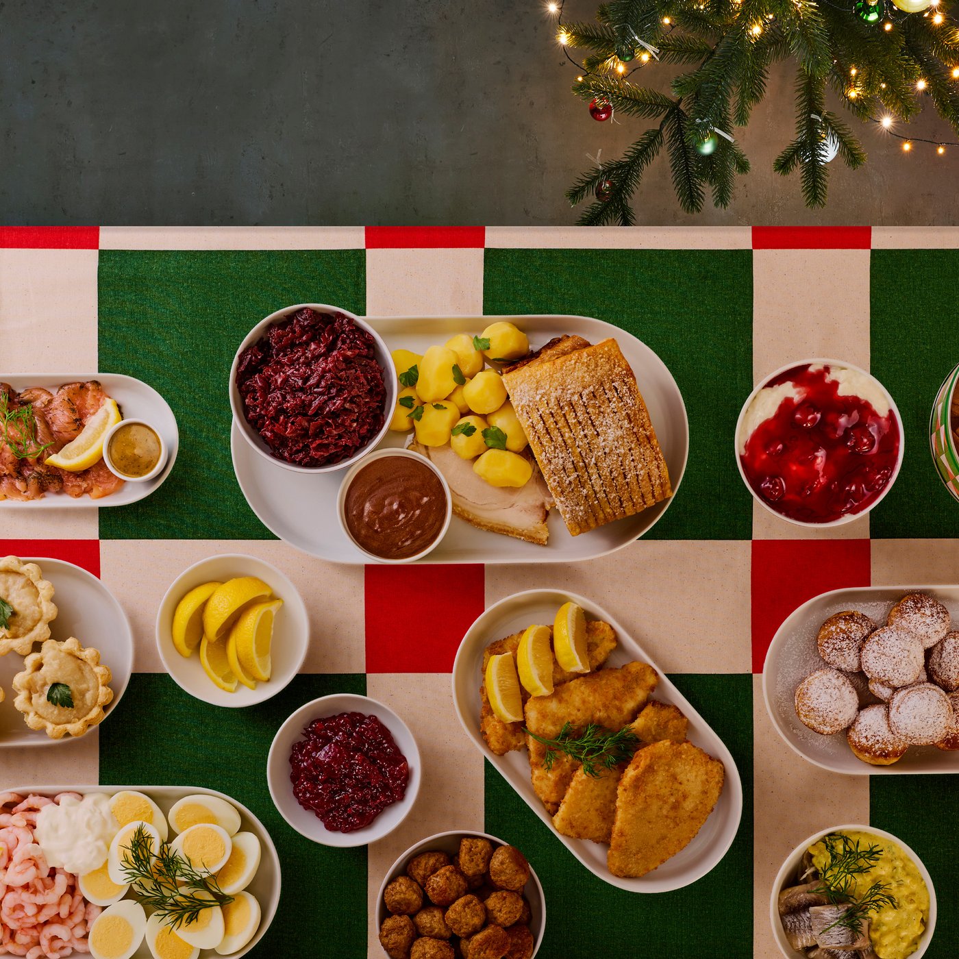Oversigtsbillede af et julebord dækket med en VINTERFINT dug i røde og grønne tern. På bordet står en masse juleretter på hver deres tallerken.