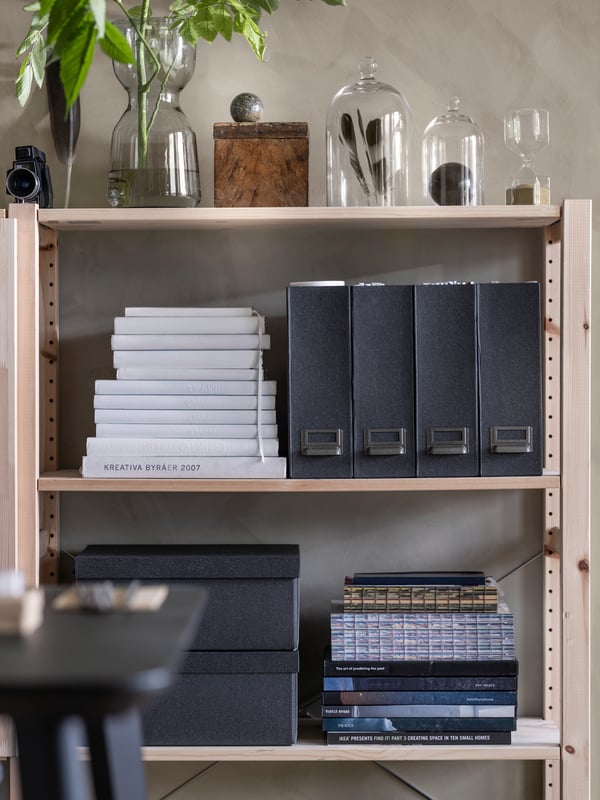 Wooden shelves against a wall with piles of books and black storage boxes and glass vases with plants and feathers inside.