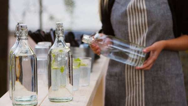 Ulkopöydällä on useita lasisia KORKEN-pulloja ja vieressä henkilö pitää yhtä niistä kädessään.