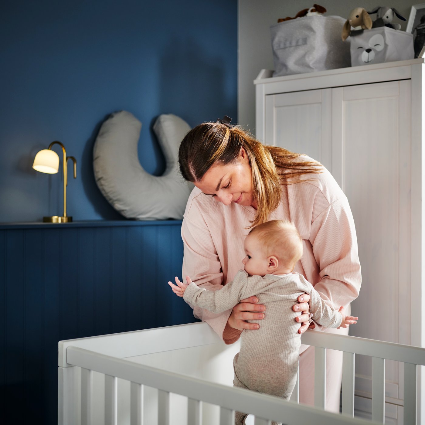 A person picks up a baby from a white SUNDVIK cot in the corner of a dark blue bedroom with a white wardrobe behind them.