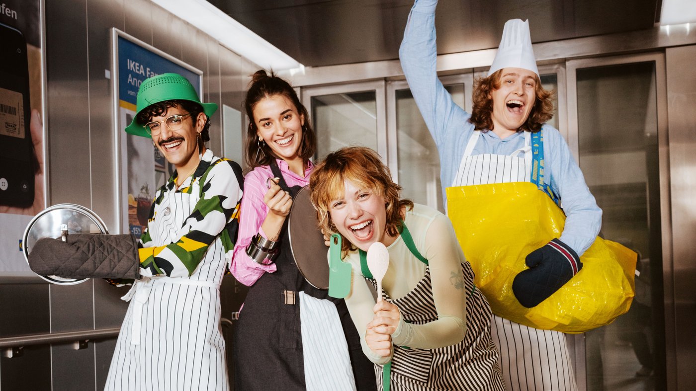 Four people standing in an IKEA elevator, wearing kitchen clothes and holding kitchen utensils.