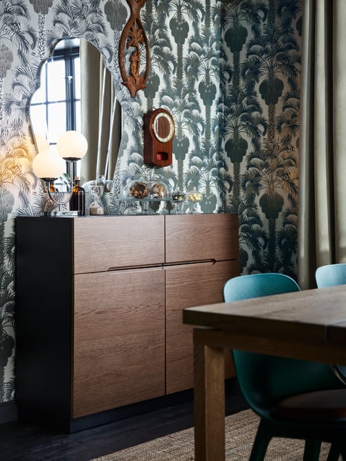 Ein KLACKENÄS Sideboard mit einem Spiegel und Holzdekorationen an der Wand darüber. Obenauf steht eine Tischleuchte. Neben dem Sideboard ist ein Esstisch zu sehen.