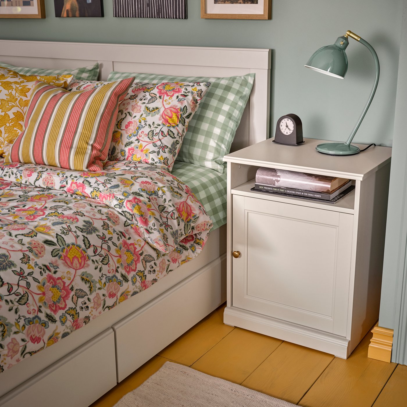 Una recámara con sábanas DAGGSALVIA sobre una cama junto a un buró color hueso FÅGELFJÄLLET con un reloj y lámpara.