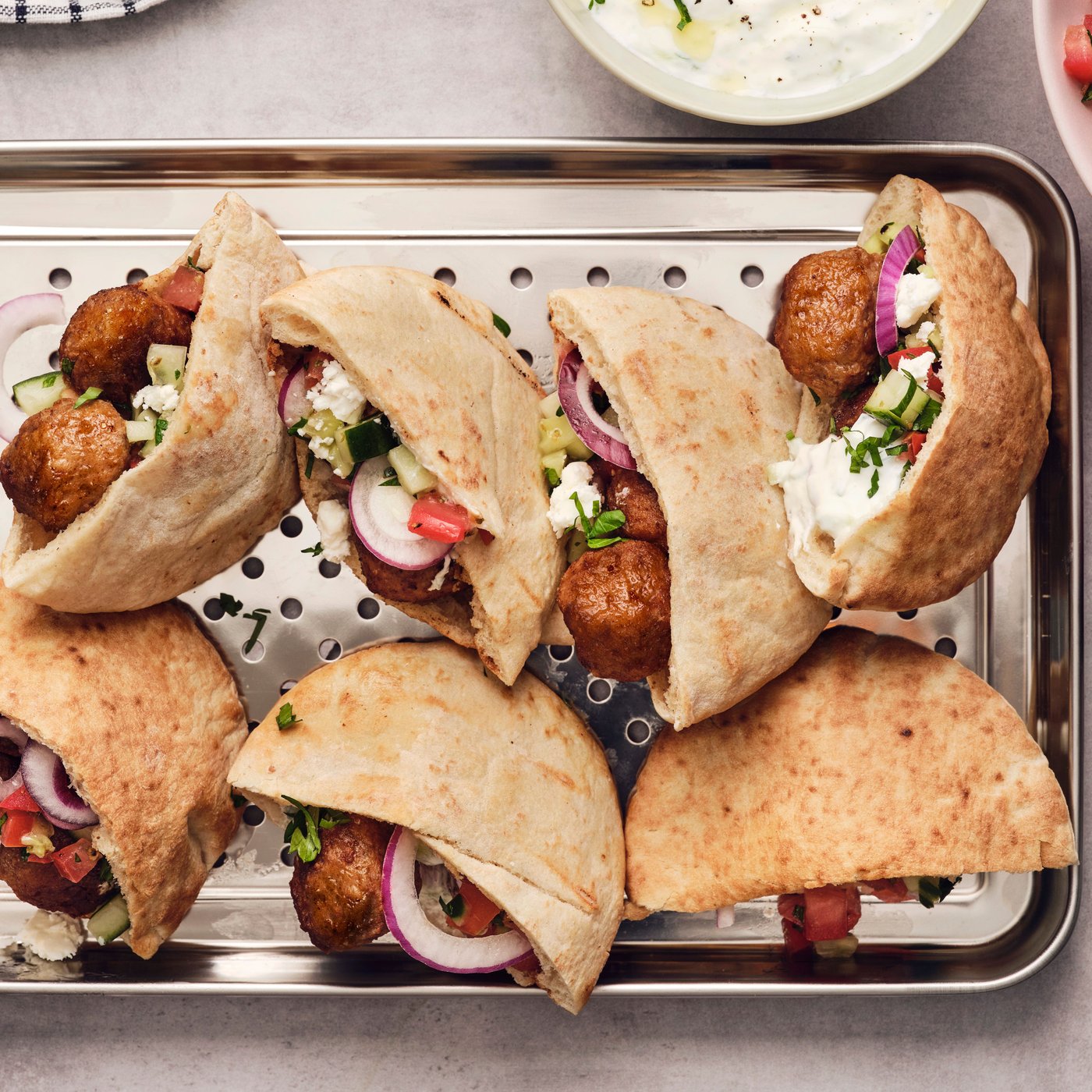 Eight pieces of pita bread are filled with meatballs and tzatziki, placed on a metal tray next to a towel and two bowls.