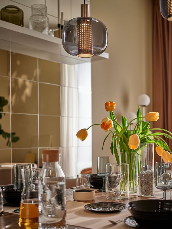 A dining table is set with black bowls and plates, glassware and vase with tulips, with HÖGVIND pendant lamps hanging above.