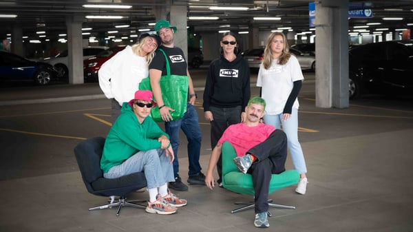 Sex personer poserar i ett parkeringsgarage, klädda i avslappnade och IKEA inspirerade outfits. Två sitter på stolar medan resten står, med bilar och betongpelare i bakgrunden.