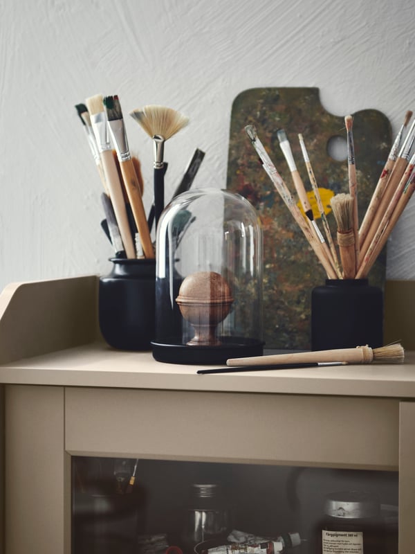 Sprawling bouquets of thin paintbrushes placed in a black KONSTFULL vase and a black jar, atop a HAUGA cabinet.