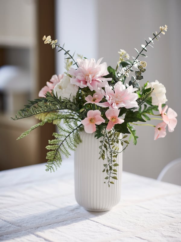 A SMYCKA artificial bouquet and some SMYCKA artificial flowers stand in a white STILREN vase on a white surface.