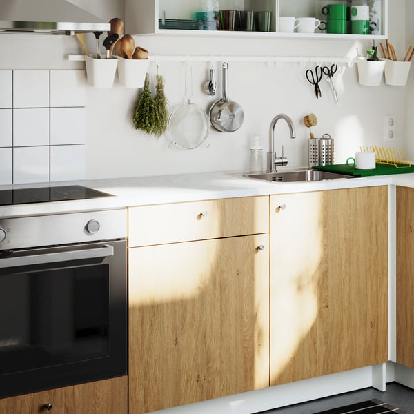 A sun-drenched KNOXHULT kitchen with fronts in oak effect. There are scissors, herbs and a pot hanging from a white rail.