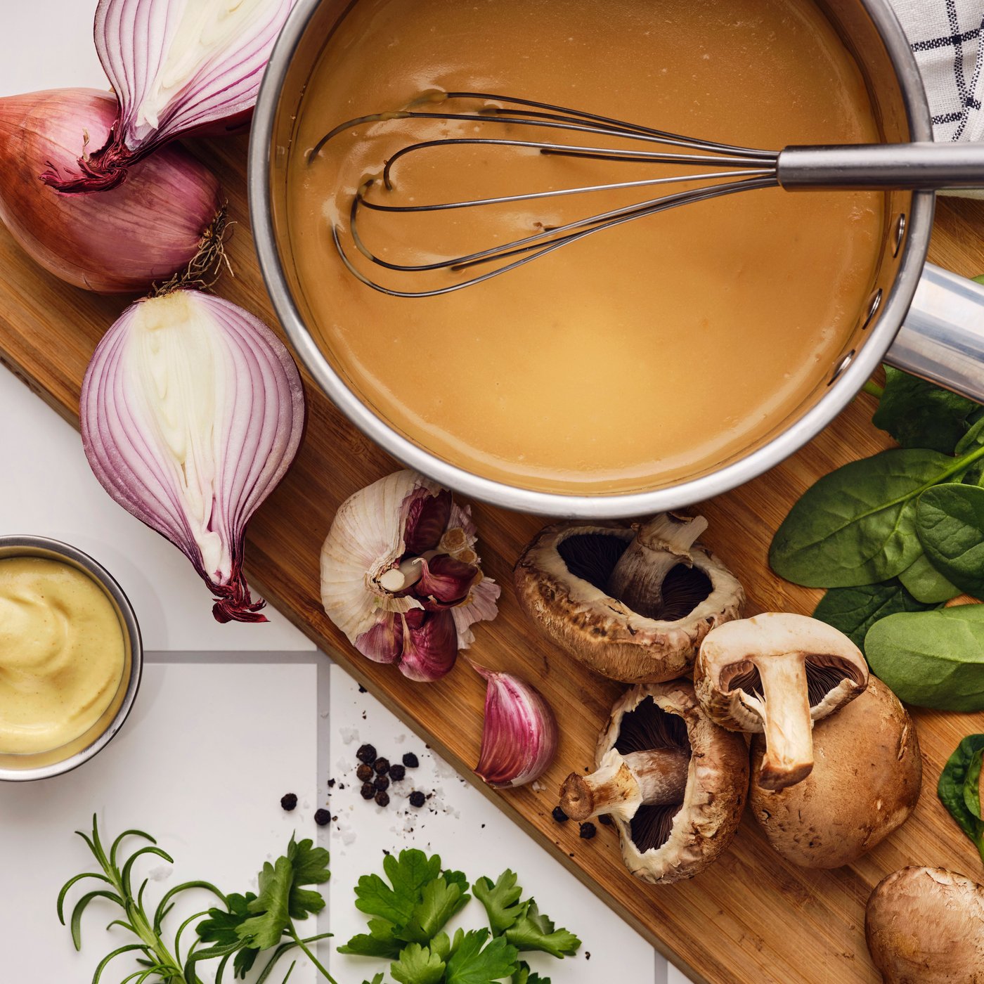 Op een snijplank liggen knoflook, champignons, kruiden en sjalotten. In een steelpan wordt met een garde ALLEMANSRÄTTEN sausmix klaargemaakt.