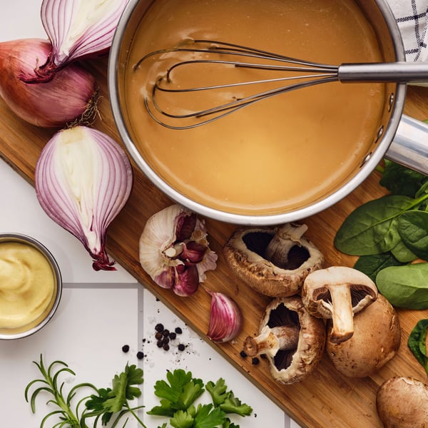 On a cutting board with garlic, mushrooms, herbs and shallots, a whisk is in a saucepan with ALLEMANSRÄTTEN savory sauce mix.