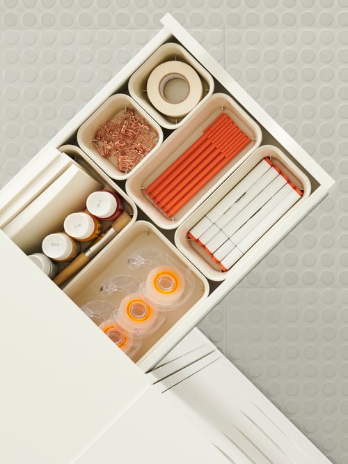 A drawer sticking out from a white unit holding different organisers with tape, pens, paper clips etc. with orange accents.