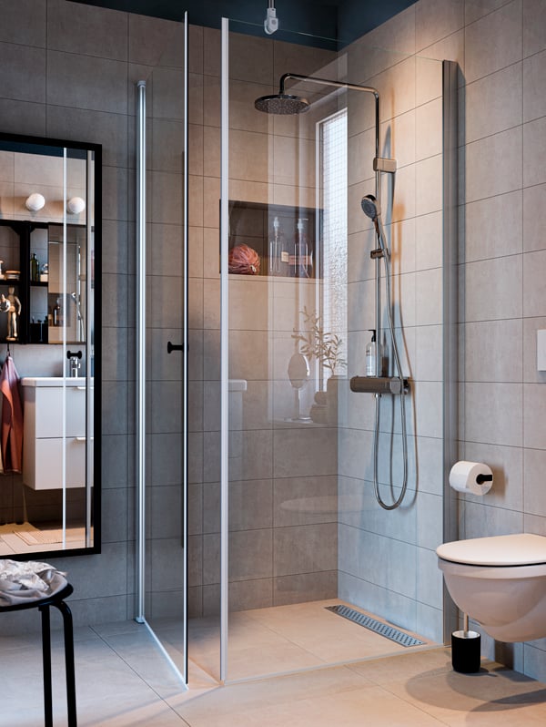 Wyłożona szarymi płytkami łazienka ze szklanymi ścianką i drzwiami kabiny prysznicowej, wysokim lustrem i chromowanym zestawem prysznicowym.