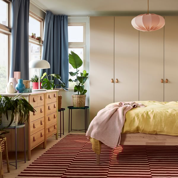 Ein graubeiger Kleiderschrank mit REINSVOLL Türen, blaue Gardinen, eine rosafarbene Hängeleuchte und gelbe Betttextilien.
