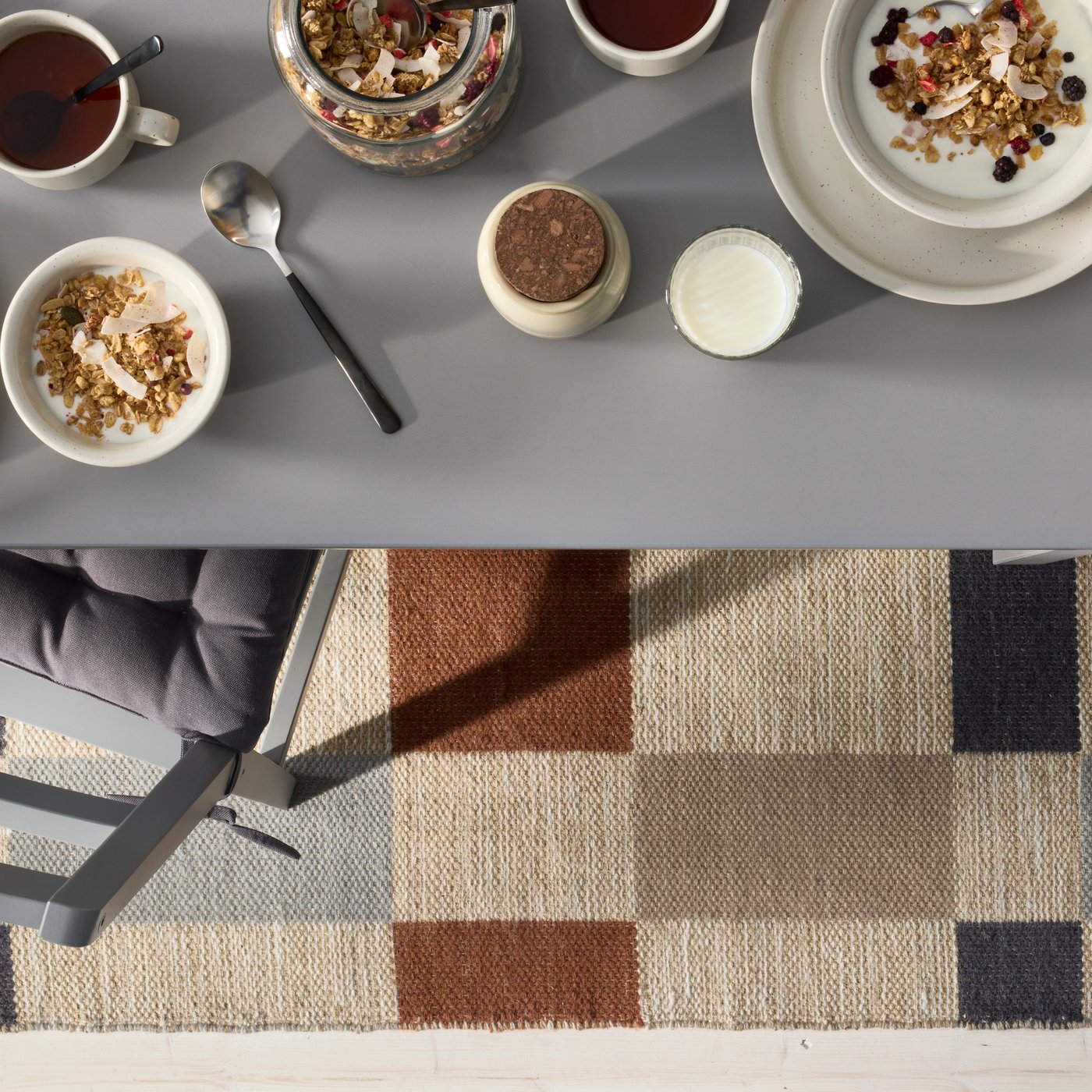 A grey/brown checkered BLÅLJUS flatwoven rug complements a dining table set with bowls, a spoon, and granola.