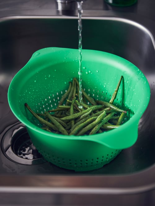 Gros plan sur un évier de cuisine avec une grande passoire en plastique vert avec poignées, contenant des haricots verts, sous le robinet.