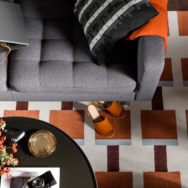 Överblick av en grå LANDSKRONA soffa bredvid en orangevit geometriskt mönstrad matta, med ett par tofflor på golvet.