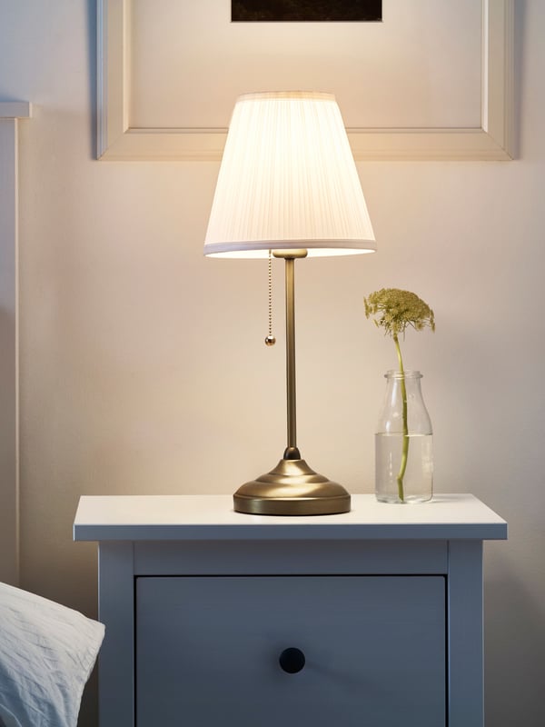 IKEA ÅRSTID brass/white table lamp on top of a bedside table in the bedroom.