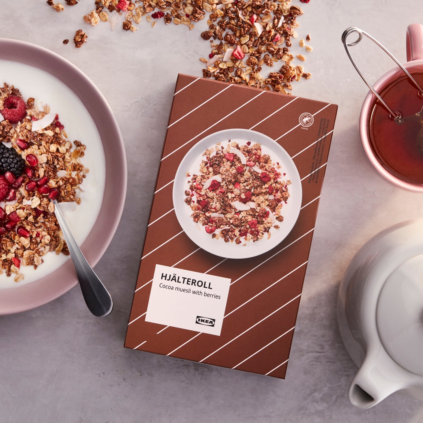 Smeđa kutija IKEA kakao musli s bobicama, uz ružičastu zdjelu jogurta i musli sa žlicom, čaj i bijeli čajnik na sivoj podlozi.
