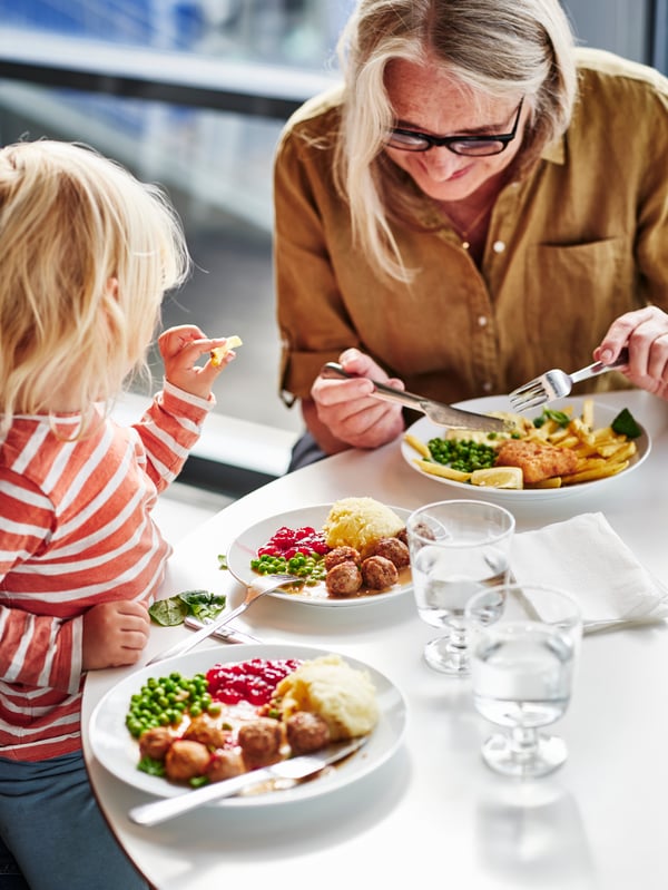 En kvinde og et barn læner sig henover et bord imens de spiser af tallerkener med kartoffelmos, ærter og tyttebærsauce.