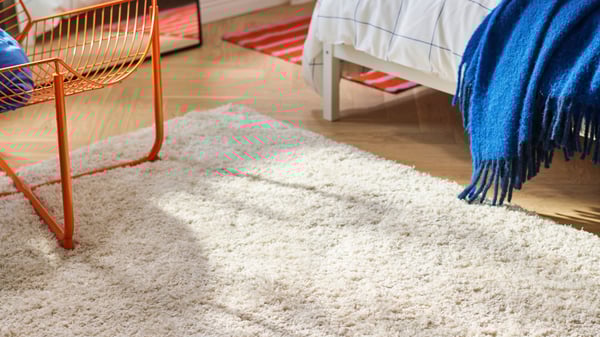 Valkoinen korkeanukkainen VOLLERSLEV-matto auringon valaisemalla lattialla sängyn vieressä.