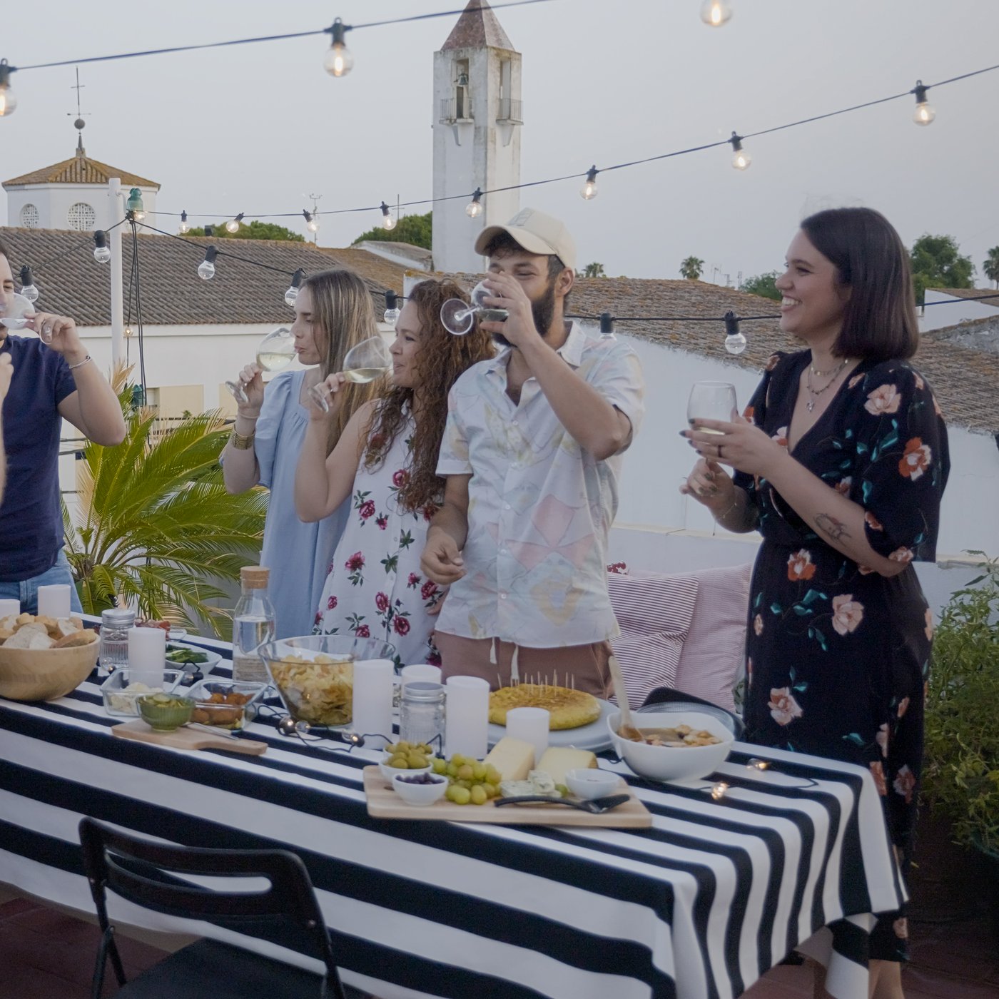 Un gruppo di amici che mangiano, bevono e conversano allegramente, in piedi accanto a un buffet ricco di pietanze, su una terrazza all'aperto.