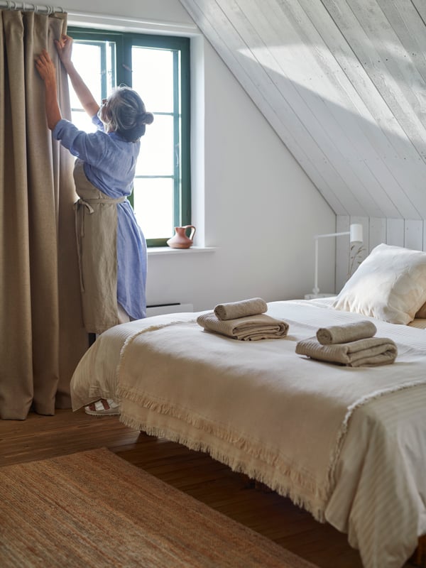 A person wearing a dress and an apron is opening the curtains in a bedroom in rural style with a slanted roof.