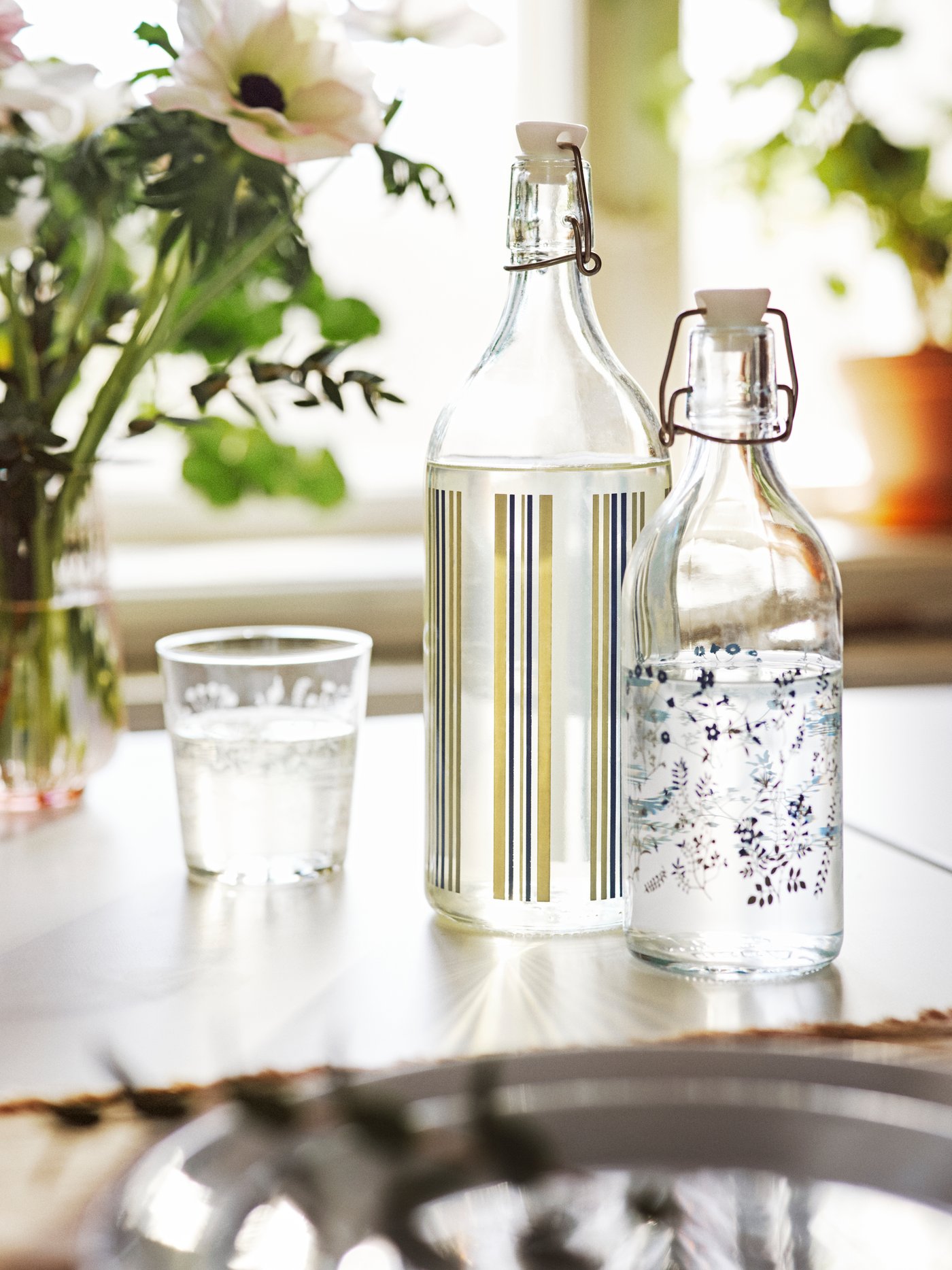 Dua botol dengan penyumbat KORKEN yang bercorak terletak di atas meja dengan gelas bercorak HAJMAL di hadapan vas berisi bunga.