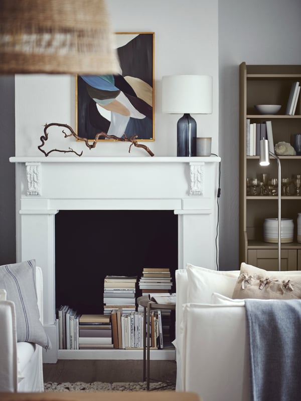 A light living room with book stacks in a decorative fireplace, a beige HAUGA high cabinet and a white BACKSÄLEN armchair.